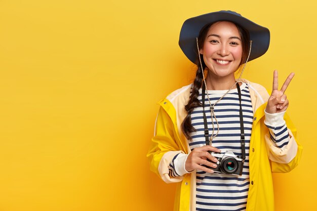 Sorridente viaggiatore femminile fa un gesto di pace, scatta foto con la fotocamera retrò, indossa un cappello, maglione a righe e impermeabile, gode di viaggi emozionanti, posa su sfondo giallo, copia spazio per il testo