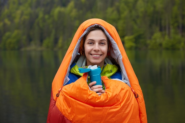 Sorridente viaggiatrice di buon umore, avvolta nel sacco a pelo, trascorre il tempo libero nella natura, tiene thermos di bevanda calda