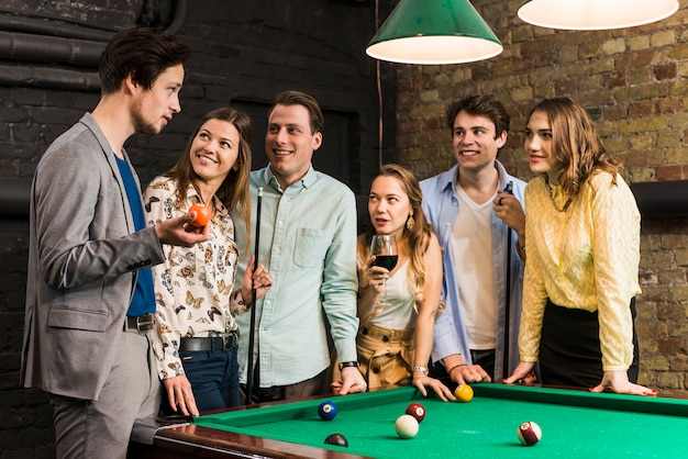 Sorridenti amici maschi e femminili guardando uomo che tiene palla da biliardo nel club