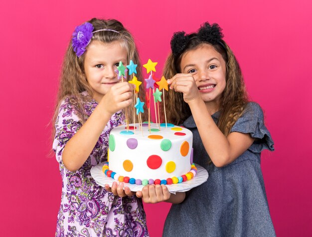 Sorridenti bambine graziose che tengono insieme la torta di compleanno isolata sulla parete rosa con lo spazio della copia