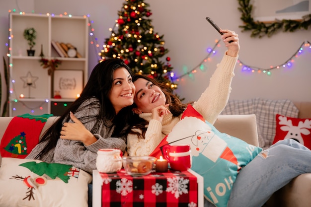 sorridenti belle ragazze prendono selfie seduti sulle poltrone e godersi il periodo natalizio a casa