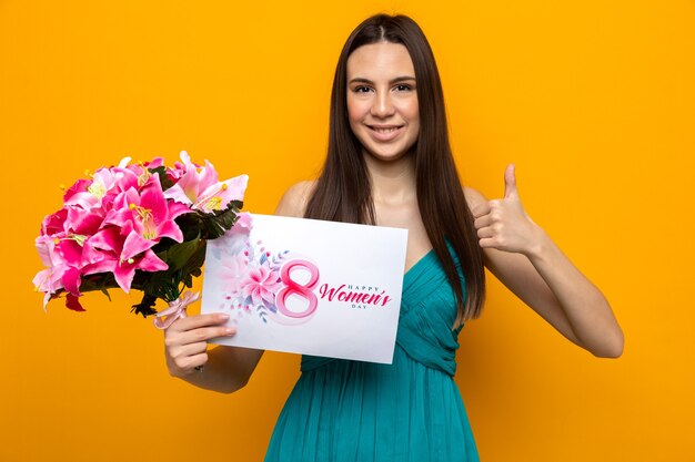 Sorridere mostrando pollice in su bella ragazza il giorno della donna felice tenendo la cartolina con bouquet isolato su parete arancione