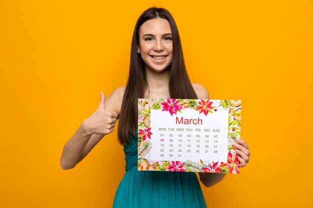 Sorridere mostrando pollice in su bella ragazza il giorno delle donne felici tenendo il calendario isolato sulla parete arancione