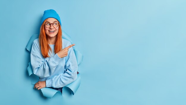 Sorrisi attraenti felici dell'adolescente ha ampiamente i capelli rossi lunghi indossa la felpa e il cappello blu.