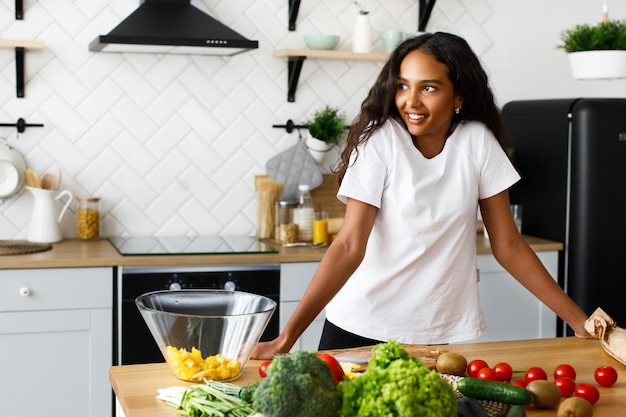 Sorriso bella mulatta è in piedi vicino al tavolo pieno di verdure fresche sulla cucina moderna