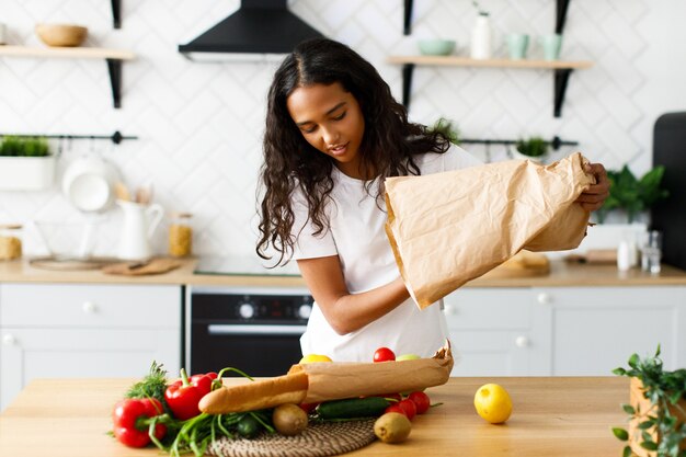 Sorriso bella mulatta sta preparando un pasto a base di verdure fresche sul tavolo della cucina moderna