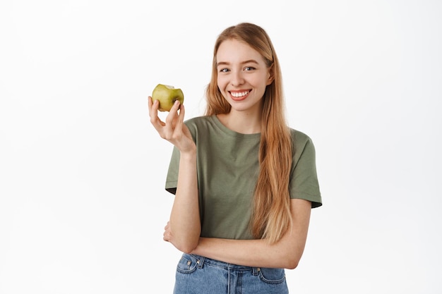 Sorriso bianco. Felice giovane donna che mangia mela verde, mostra denti perfetti e sembra soddisfatta della telecamera, in piedi in maglietta su sfondo bianco