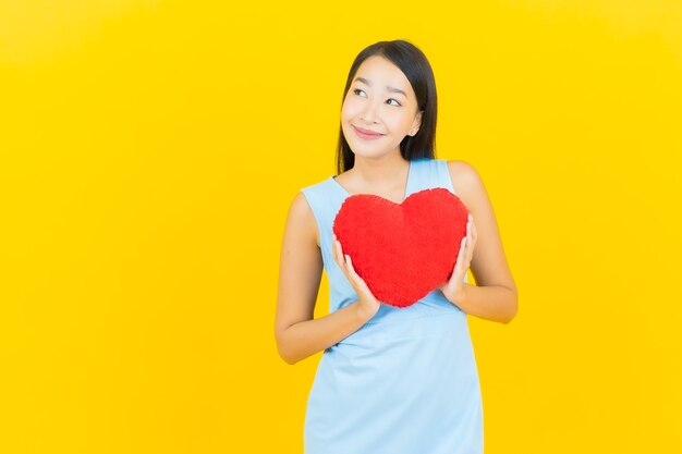 Sorriso della bella giovane donna asiatica del ritratto con forma del cuscino del cuore sulla parete di colore giallo
