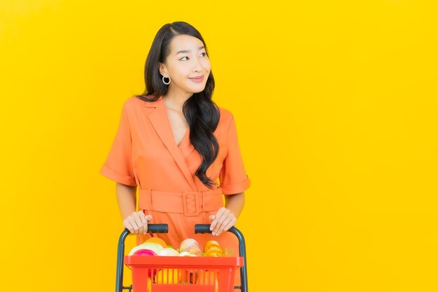 Sorriso della bella giovane donna asiatica del ritratto con il cestino della drogheria dal supermercato su colore giallo