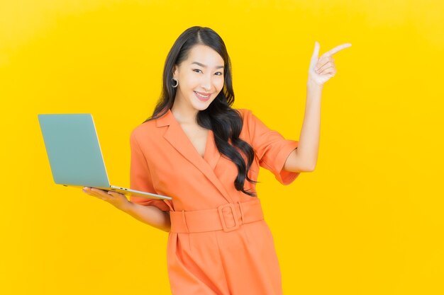 Sorriso della bella giovane donna asiatica del ritratto con il computer portatile del computer su colore giallo