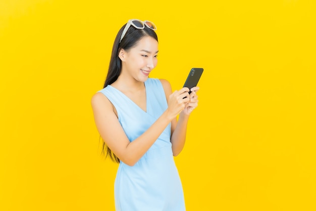 Sorriso della bella giovane donna asiatica del ritratto con il telefono cellulare astuto sulla parete di colore giallo