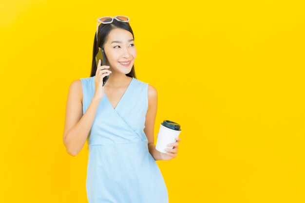 Sorriso della bella giovane donna asiatica del ritratto con il telefono cellulare astuto sulla parete di colore giallo
