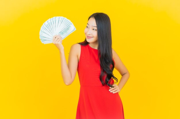 Sorriso della bella giovane donna asiatica del ritratto con molti contanti e soldi sulla parete gialla