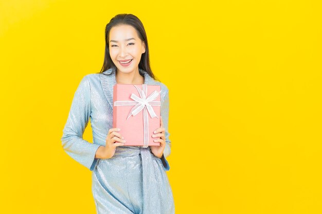 Sorriso di bella giovane donna asiatica del ritratto con confezione regalo rossa sulla parete gialla