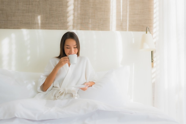 Sorriso felice della bella giovane donna asiatica del ritratto con la tazza di caffè sul letto