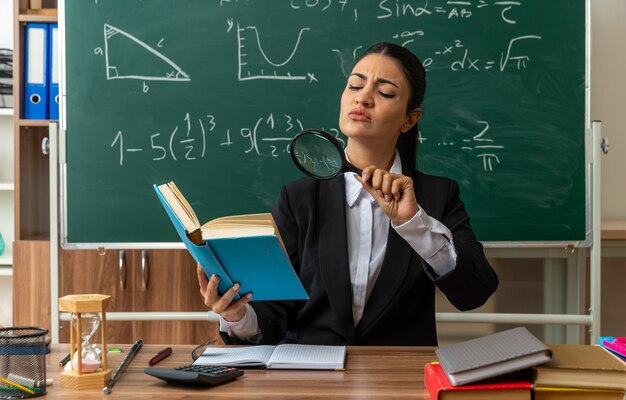 sospettosa giovane insegnante femminile si siede al tavolo con materiale scolastico libro di lettura con lente d'ingrandimento in classe