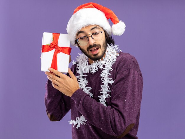 Sospettoso giovane bel ragazzo che indossa il cappello di Natale con la ghirlanda sul collo che tiene e ascolta confezione regalo isolato su sfondo blu