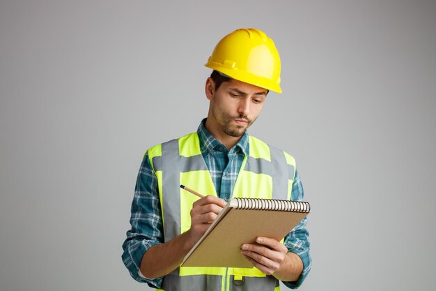 Sospettoso giovane ingegnere maschio che indossa un casco di sicurezza e uniforme che tiene blocco note e matita che prende appunti di lavoro isolati su sfondo bianco