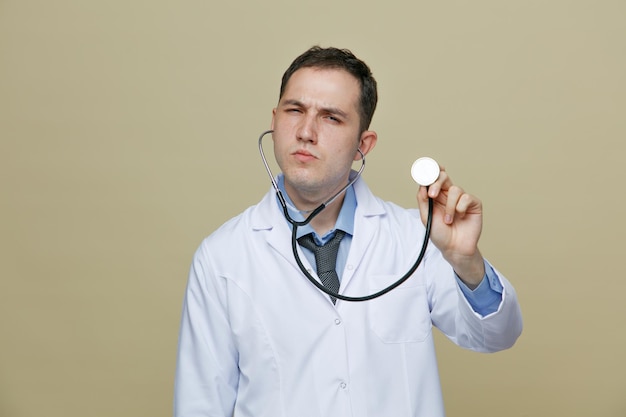 Sospettoso giovane medico maschio che indossa una veste medica e uno stetoscopio guardando la fotocamera che afferra uno stetoscopio che lo mostra isolato su sfondo verde oliva
