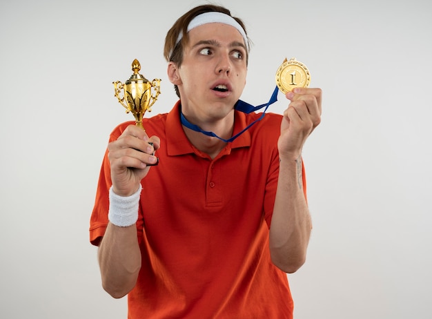 Sospettoso giovane ragazzo sportivo che indossa la fascia con il braccialetto che tiene la tazza del vincitore con la medaglia isolata sulla parete bianca