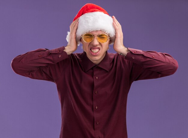 Sottolineato giovane uomo biondo che indossa santa cappello e occhiali tenendo le mani sulla testa con gli occhi ben chiusi isolati sulla parete viola