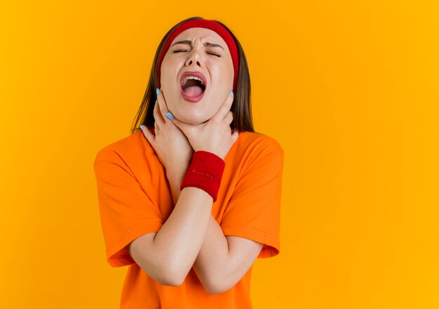 Sottolineato stufo giovane donna sportiva che indossa fascia e braccialetti che si soffoca con gli occhi chiusi isolati sulla parete arancione con spazio di copia