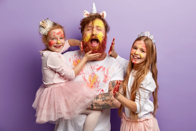 Sovraccarico di stanchezza padre single con la barba rossa, piange disperatamente, si diverte con due bambine, usa vernici colorate, ha espressioni felici, sta sul muro viola. Concetto di giorno di padri felice