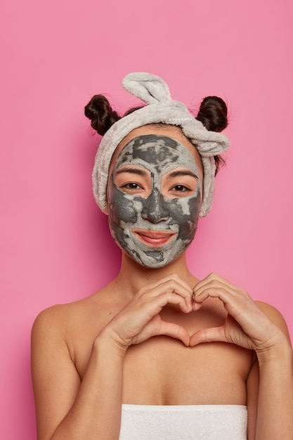 Spa donna etnica indossa una maschera di bellezza sul viso, forma il cuore sul corpo, esprime amore, fa procedure anti rughe dopo aver fatto il bagno, isolato su un muro roseo. Concetto di bellezza e benessere