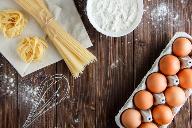 Spaghetti con uova, farina, frusta, fettuccine su asciugamano di legno e cucina, piatto disteso.