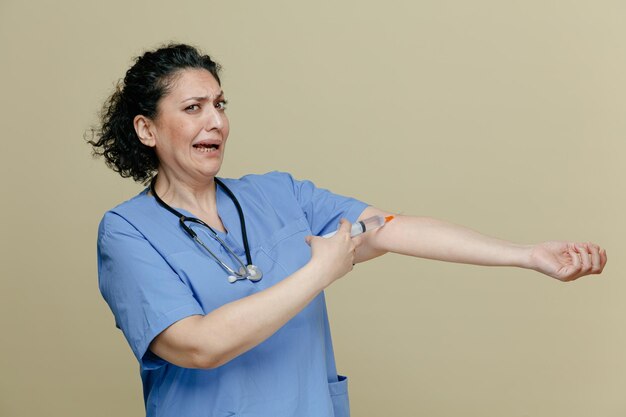 Spaventata dottoressa di mezza età che indossa uniforme e stetoscopio intorno al collo guardando la telecamera allungando la mano dando iniezione nella sua stessa vena isolata su sfondo verde oliva