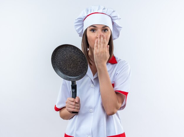 Spaventata giovane cuoca che indossa l'uniforme dello chef tenendo la padella coperta faccia con la mano isolata su sfondo bianco