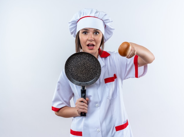 Spaventata giovane cuoca che indossa l'uniforme dello chef tenendo la padella e tenendo fuori il cucchiaio in telecamera isolata su sfondo bianco