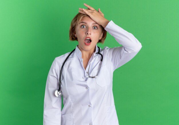 Spaventata giovane dottoressa che indossa un abito medico con uno stetoscopio che mette la mano sulla fronte isolata su sfondo verde