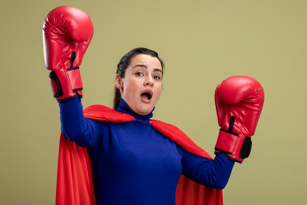 Spaventata giovane ragazza del supereroe che indossa i guantoni da boxe alzando le mani isolato su sfondo verde oliva