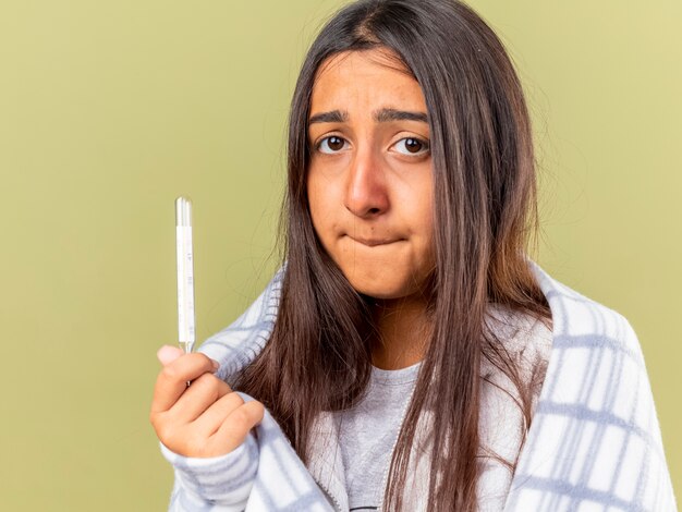 Spaventata giovane ragazza malata avvolta in plaid che tiene il termometro morde le labbra isolate su verde oliva