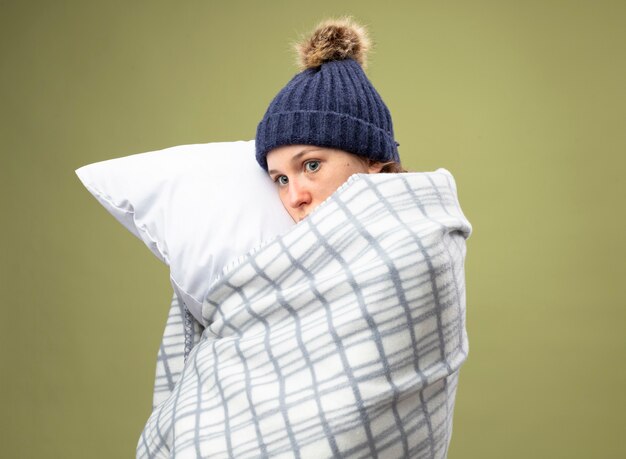 Spaventata giovane ragazza malata guardando al lato che indossa una veste bianca e cappello invernale con sciarpa avvolta in un plaid abbracciato cuscino