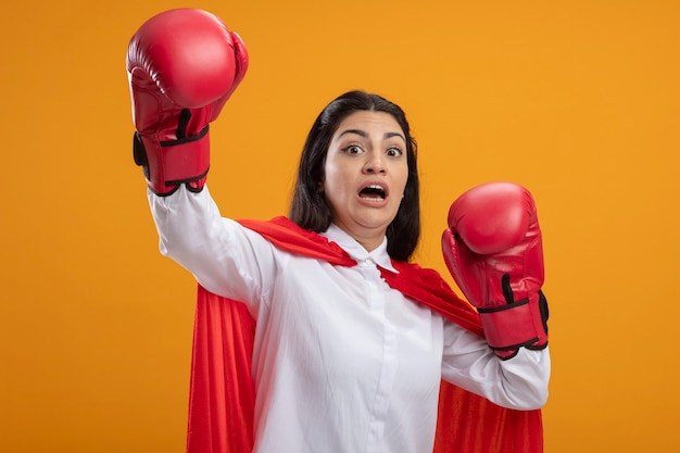 Spaventata giovane superdonna che indossa guanti box guardando davanti alzando le mani in alto isolato sulla parete arancione