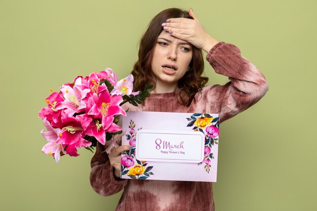 Spaventata mettendo la mano sulla fronte bella ragazza in felice festa della donna che tiene bouquet con biglietto di auguri