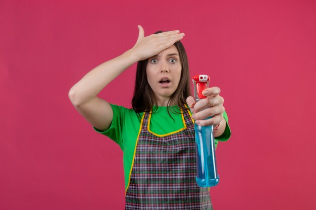 Spaventata pulizia ragazza giovane indossa uniforme che tiene spray per la pulizia mise la mano sulla fronte isolato su sfondo rosa