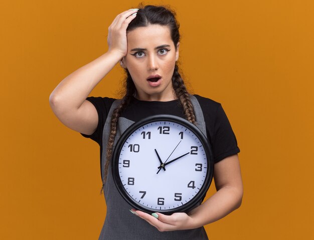 Spaventato giovane barbiere femminile in uniforme che tiene l'orologio da parete e mettendo la mano sulla testa isolata sulla parete arancione