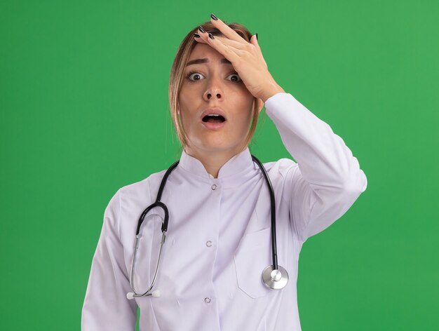 Spaventato giovane medico femminile che indossa abito medico con lo stetoscopio che mette la mano sulla fronte isolata sulla parete verde
