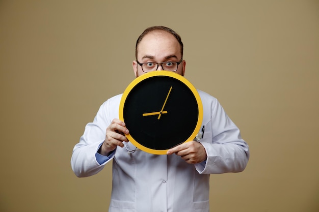 Spaventato giovane medico maschio che indossa occhiali camice da laboratorio e stetoscopio intorno al collo tenendo l'orologio nascosto dietro di esso guardando la fotocamera da dietro l'orologio isolato su sfondo verde oliva