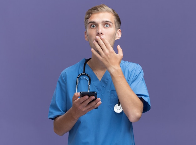 Spaventato giovane medico maschio indossa uniforme medico con lo stetoscopio che tiene il telefono e la bocca coperta con la mano isolata sulla parete blu