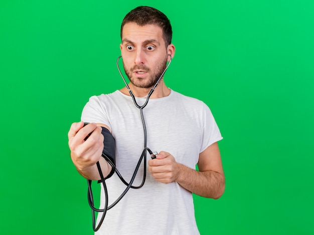 Spaventato giovane uomo malato che misura la propria pressione con sfigmomanometro isolato su sfondo verde