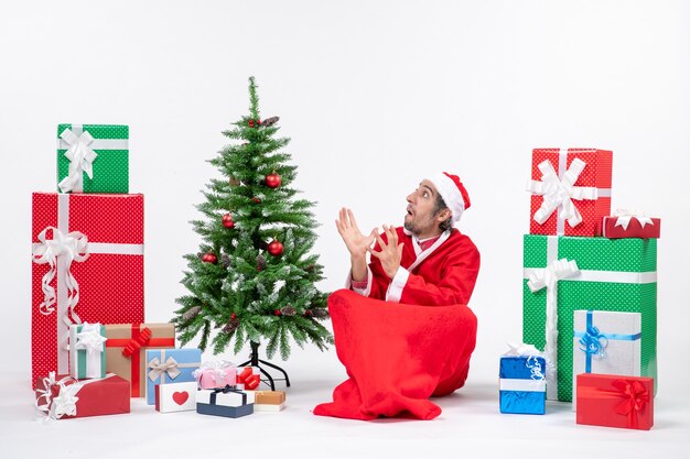 Spaventato il giovane adulto vestito da Babbo Natale con doni e albero di Natale decorato seduto per terra su sfondo bianco