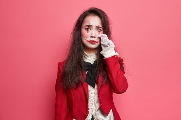 Spaventosa donna bruna sconsolata con il trucco di halloween salviette le lacrime ha un'espressione cupa faccia insanguinata l'arte indossa giacca rossa e guanti di pizzo lente bianca sull'occhio pone al coperto contro il muro roseo