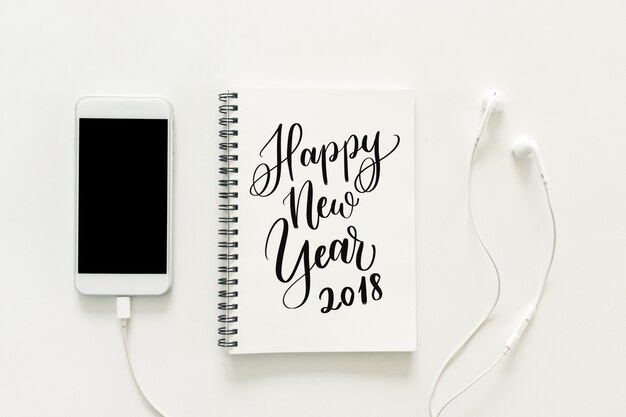 Spazio minimo di lavoro - foto piatta piano creativo della scrivania di lavoro con &quot;2018 felice anno nuovo&quot; sul libro di schizzi e telefono cellulare con schermo vuoto e auricolare su sfondo bianco. Vista dall&#39;alto, concetto di nuovo anno.