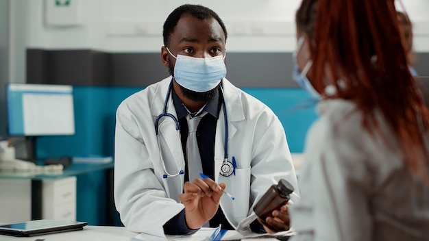 Specialista della maternità e donna incinta con maschera facciale alla visita di controllo, incontro per parlare di gravidanza. Futura madre con pancione che chiede consiglio medico al medico alla scrivania del gabinetto.