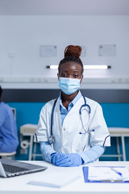 Specialista medico ospedaliero seduto al tavolo dell'ufficio mentre indossa una maschera protettiva contro i virus e guanti chirurgici. Esperto di assistenza sanitaria clinica seduto alla scrivania dell'ufficio mentre indossa la maschera facciale