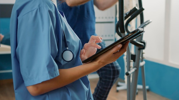 Specialista sanitario in uniforme che utilizza tablet digitale per aiutare il paziente anziano con lesioni fisiche e disturbi meccanici. Dispositivo di sostegno dell'infermiera durante la procedura di riabilitazione muscolare. Avvicinamento.